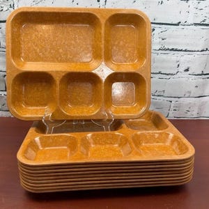 May include: A stack of rectangular amber-colored plastic cafeteria trays, each featuring five distinct compartments of varying sizes. The trays have a mottled, textured finish and are arranged with one tray standing upright on a clear stand.