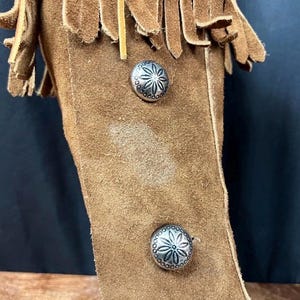 May include: Close-up of a brown suede leather item with decorative silver metal buttons. The item has a fringe detail at the top. The buttons have a floral design.