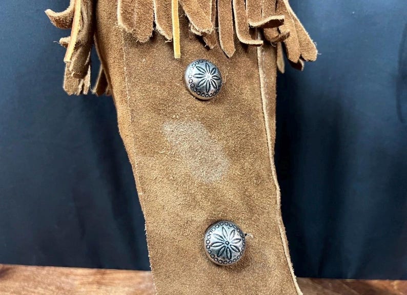 May include: Close-up of a brown suede leather item with decorative silver metal buttons. The item has a fringe detail at the top. The buttons have a floral design.