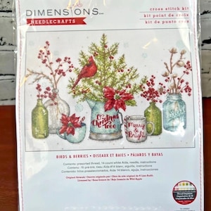 May include: A Dimensions Needlecrafts cross stitch kit titled Birds & Berries featuring a holiday scene with a cardinal, a Christmas tree in a pitcher, poinsettias, and jars. The package includes 14 count white Aida, thread, and instructions.
