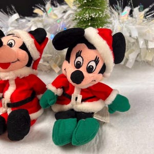 K&ouml;nnte beinhalten: Zwei Pl&uuml;schfiguren von Mickey Mouse in Weihnachtsmann-Outfits. Jede Figur tr&auml;gt eine rote Jacke mit wei&szlig;em Besatz, einen schwarzen G&uuml;rtel, gr&uuml;ne Handschuhe und Schuhe sowie eine Weihnachtsm&uuml;tze. Der Hintergrund umfasst einen kleinen Weihnachtsbaum und wei&szlig;en Lametta.