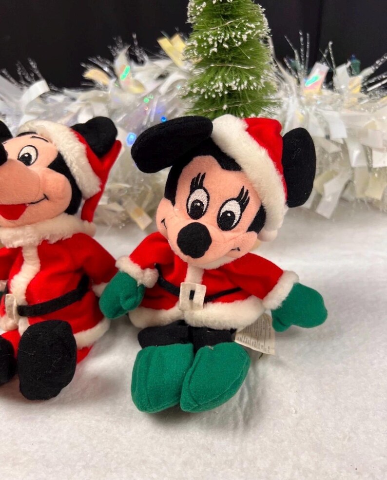 K&ouml;nnte beinhalten: Zwei Pl&uuml;schfiguren von Mickey Mouse in Weihnachtsmann-Outfits. Jede Figur tr&auml;gt eine rote Jacke mit wei&szlig;em Besatz, einen schwarzen G&uuml;rtel, gr&uuml;ne Handschuhe und Schuhe sowie eine Weihnachtsm&uuml;tze. Der Hintergrund umfasst einen kleinen Weihnachtsbaum und wei&szlig;en Lametta.