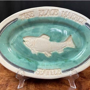 May include: Oval ceramic serving dish with a teal interior and a cream-colored exterior. The words "PIKE PLACE MARKET" and "SEATTLE" are embossed on the rim. A fish is embossed in the center.