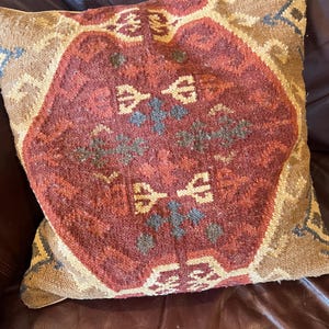 May include: Square decorative pillow with a geometric pattern in shades of red, brown, and beige. The pillow features a central medallion design with symmetrical motifs and a textured appearance, suitable for home decor.