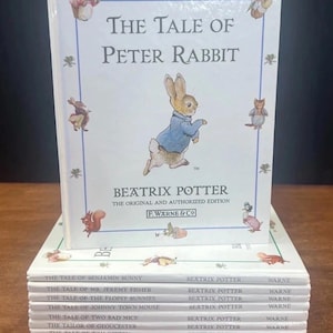 May include: A stack of white hardcover books by Beatrix Potter. The top book is The Tale of Peter Rabbit, featuring a central illustration of a rabbit in a blue jacket. Below it are nine other titles including The Tale of Benjamin Bunny and others.
