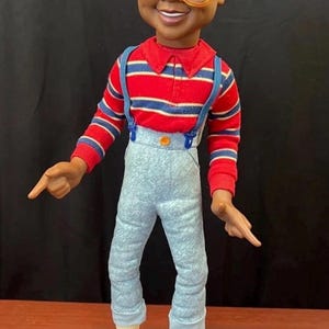May include: A plastic doll with a dark complexion, short black hair, and large orange-rimmed glasses. The doll wears a red, blue, and white striped long-sleeve shirt, light blue textured pants with blue suspenders, white socks, and yellow shoes.
