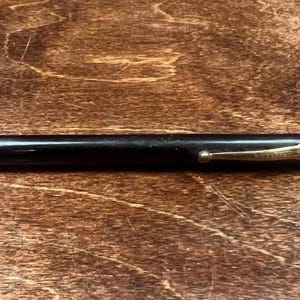 Op de afbeelding: Een zwarte mechanische potlood met een goudkleurige punt en accenten. De potlood heeft een slank, gestroomlijnd ontwerp en staat tegen een houten achtergrond. Op de clip van de potlood staat "SHEAFFER'S".