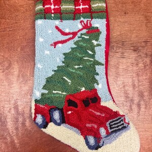 Puede incluir: Calcetín navideño hecho a mano con un camión rojo que transporta un árbol de Navidad verde. El calcetín tiene un fondo azul con acentos de nieve blanca y una parte superior a cuadros rojos. Un lazo rojo está en la parte superior para colgar.