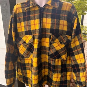 May include: A yellow and black plaid flannel shirt with a half-zip closure and two front pockets. The shirt has long sleeves and a collared neckline. The plaid pattern includes red accent lines. The shirt is displayed on a mannequin.