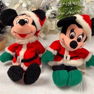K&ouml;nnte beinhalten: Zwei Pl&uuml;schfiguren von Mickey und Minnie Mouse in Weihnachtsmann-Outfits. Jede Figur tr&auml;gt eine rote Jacke mit wei&szlig;em Besatz, einen schwarzen G&uuml;rtel, gr&uuml;ne Handschuhe und einen Weihnachtsmannhut. Die Figuren sind schwarz, rot, wei&szlig; und gr&uuml;n.