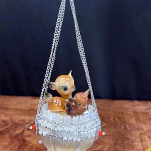 Puede incluir: Adorno navideño vintage con tres figuras de ciervos en una cesta decorativa. La cesta está adornada con cuentas blancas y adornos plateados, suspendida por una cadena plateada. Los acentos rojos y blancos añaden un toque festivo a esta decoración.