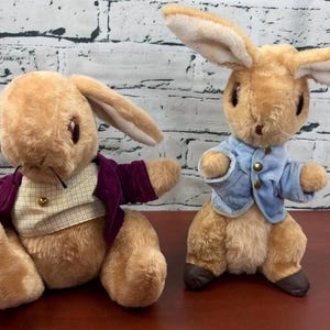 May include: Two plush rabbit toys with tan fur. The left rabbit wears a purple velvet jacket over a cream and tan checkered vest. The right rabbit stands upright, wearing a light blue velvet jacket with small gold buttons and dark brown feet.