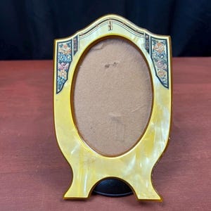 May include: An oval-shaped picture frame with a cream-colored, marbled finish. The frame features decorative accents on the upper corners with floral patterns in shades of pink, orange, and blue, set against a dark background with gold-toned lines.