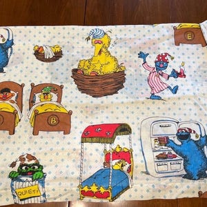 Könnte beinhalten: Weißes Sesame Street-Bettlaken mit verschiedenen Charakteren in einem sich wiederholenden Muster. Big Bird befindet sich in einem Nest, Cookie Monster an einem Kühlschrank und andere Charaktere in Betten. Das Laken hat ein hellblaues Rautenmuster.