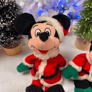 K&ouml;nnte beinhalten: Ein Pl&uuml;schtier von Mickey Mouse in einem Weihnachtsmann-Outfit. Das Spielzeug hat schwarze Ohren, einen rot-wei&szlig;en Weihnachtsmannhut, einen roten Mantel mit wei&szlig;em Besatz, gr&uuml;ne Handschuhe und schwarze Schuhe. Weihnachtsdekoration im Hintergrund.