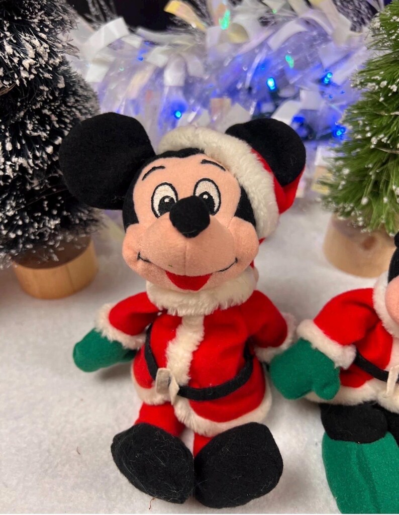 K&ouml;nnte beinhalten: Ein Pl&uuml;schtier von Mickey Mouse in einem Weihnachtsmann-Outfit. Das Spielzeug hat schwarze Ohren, einen rot-wei&szlig;en Weihnachtsmannhut, einen roten Mantel mit wei&szlig;em Besatz, gr&uuml;ne Handschuhe und schwarze Schuhe. Weihnachtsdekoration im Hintergrund.