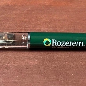 Op de afbeelding: Een groene en doorzichtige balpen met een zilveren punt en een transparant gedeelte in de houder met kleine gele zwevende elementen. De groene houder toont de witte tekst Rozerem met een cirkelvormig logo met blauwe en gele kleurverlopen.