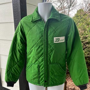 May include: Green quilted jacket with a diamond pattern, a collar, and a zipper closure. The jacket has a small pocket on the lower left side and a patch with the text "Burlington Northern" on the right chest. The cuffs are black.