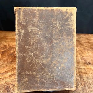 以下が含まれることがあります： 使い古された茶色の革の表紙が付いた、長方形のヴィンテージ本。表紙には、傷や風合いのある質感など、経年劣化の兆候が見られます。本の端はわずかに丸みを帯びており、アンティーク品であることを示唆しています。