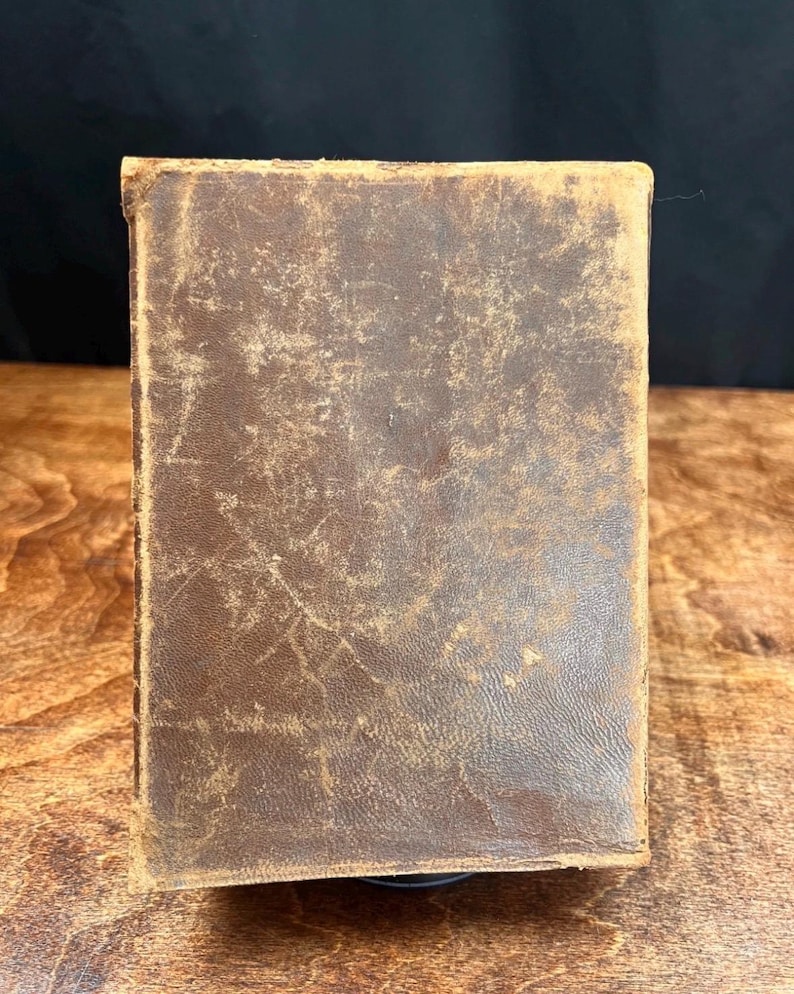 以下が含まれることがあります： 使い古された茶色の革の表紙が付いた、長方形のヴィンテージ本。表紙には、傷や風合いのある質感など、経年劣化の兆候が見られます。本の端はわずかに丸みを帯びており、アンティーク品であることを示唆しています。