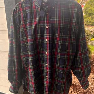 Puede incluir: Una camisa abotonada de manga larga con un patrón de cuadros clásico. La camisa tiene una base azul marino con líneas rojas, verdes y amarillas. Tiene un cuello abotonado y un bolsillo en el pecho.