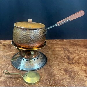 May include: A copper-colored fondue pot with a wooden handle and a small brass ladle. The pot sits on a metal stand with a burner. The pot has a hammered texture and a lid with a wooden knob.