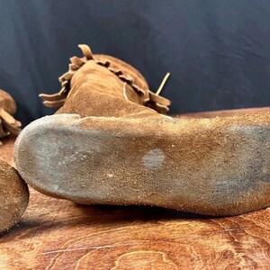 May include: Close-up of brown suede moccasins with fringed details. The shoes are made of a soft, textured material and appear to be well-worn. The soles show signs of use, suggesting they are vintage or have been used.