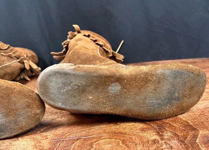 May include: Close-up of brown suede moccasins with fringed details. The shoes are made of a soft, textured material and appear to be well-worn. The soles show signs of use, suggesting they are vintage or have been used.