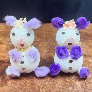 May include: Two decorative white Easter bunny figurines. Each bunny has purple ears, paws, and feet, with a small flower crown. They have black eyes, a red heart-shaped nose, and a gold bow tie. The bunnies are sitting on a wooden surface.
