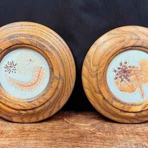 May include: Two round wooden frames with botanical art. Each frame has a light blue background with pressed leaves and flowers in shades of orange and brown. The frames are made of light brown wood.