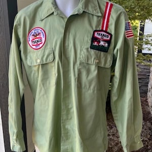 May include: Light green button-down shirt with two chest pockets. Features Texaco and National Truck Roadeo 1971 patches, plus a small American flag. The shirt has long sleeves and a collared neckline.