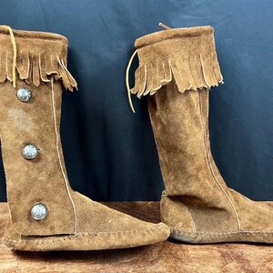 May include: A pair of knee-high brown suede boots with fringe trim at the top. The boots have decorative silver buttons and a moccasin-style sole. The boots are made of soft suede material.