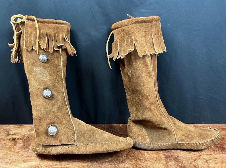 May include: A pair of knee-high brown suede boots with fringe trim at the top. The boots have decorative silver buttons and a moccasin-style sole. The boots are made of soft suede material.