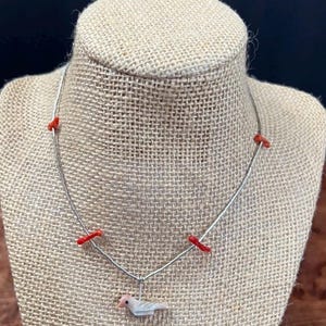 May include: A silver necklace with a bird pendant. The necklace has red coral accents and a small bird charm with pink and white details. The necklace is displayed on a burlap-covered mannequin.
