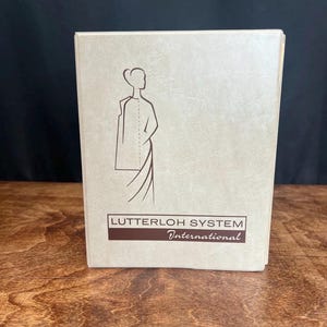 May include: A tan-colored book with the text "Lutterloh System International" in a brown rectangle. The cover features a line drawing of a person wearing a dress. The book is on a wooden surface.