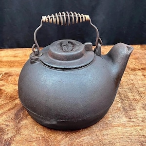 Peut inclure: Une théière en fonte brun foncé avec une poignée en fil de fer et un bec verseur. La poignée est faite d'un fil enroulé. Le couvercle a un motif en relief. La théière est posée sur une surface en bois.