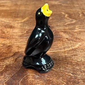 May include: A glossy black ceramic bird figurine with a bright yellow beak and eye details. The bird is perched on a base with embossed text. The figurine has a smooth, reflective surface.