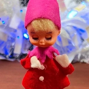 May include: A small doll with blonde hair, wearing a bright pink pointed felt hat, a red felt coat with a pink collar, a single white dot on the front, white felt mittens, and white felt boots, featuring painted eyes with long black eyelashes.