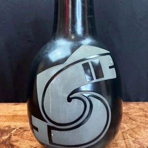 May include: Black ceramic vase with a glossy finish. The vase features a gray, stylized design on the front. The design appears to be an abstract representation of a wave and geometric shapes. The vase has a rounded body and a narrow neck.