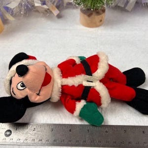 K&ouml;nnte beinhalten: Ein Pl&uuml;sch-Mickey Mouse als Weihnachtsmann, tr&auml;gt einen roten Anzug mit wei&szlig;em Besatz, einen schwarzen G&uuml;rtel und eine Weihnachtsm&uuml;tze. Das Spielzeug hat schwarze Ohren und F&uuml;&szlig;e sowie gr&uuml;ne Handschuhe. Die L&auml;nge betr&auml;gt ca. 15 cm.