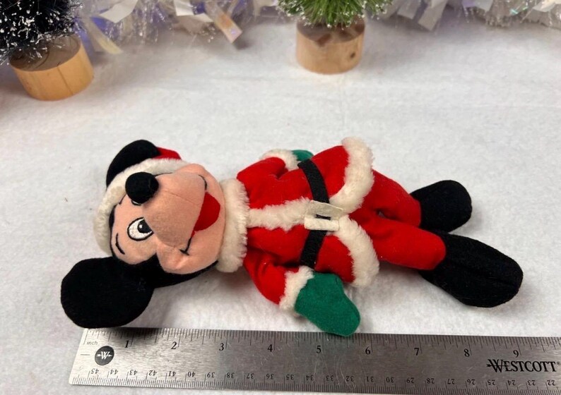 K&ouml;nnte beinhalten: Ein Pl&uuml;sch-Mickey Mouse als Weihnachtsmann, tr&auml;gt einen roten Anzug mit wei&szlig;em Besatz, einen schwarzen G&uuml;rtel und eine Weihnachtsm&uuml;tze. Das Spielzeug hat schwarze Ohren und F&uuml;&szlig;e sowie gr&uuml;ne Handschuhe. Die L&auml;nge betr&auml;gt ca. 15 cm.