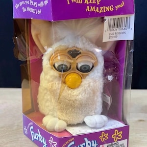 May include: A white electronic Furby toy with large eyes and a yellow beak, packaged in a purple box. The box features the text "I will KEEP AMAZING you!" and "The more you PLAY with me the MORE I do!" along with a barcode and warning label.