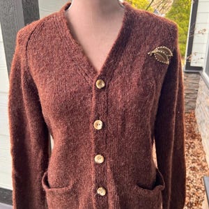 Könnte beinhalten: Ein brauner V-Ausschnitt-Cardigan mit fünf beigefarbenen Knöpfen und zwei Vordertaschen. Eine goldfarbene Blattbrosche ist auf der linken Schulter angesteckt. Der Pullover ist aus einem strukturierten Strickmaterial.