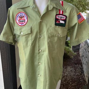 May include: A light olive green short-sleeve button-down shirt featuring two chest pockets. Patches include a round 1979 National Truck Roadeo Texaco emblem, a rectangular Texaco logo patch with a red and white stripe, and a United States flag patch.
