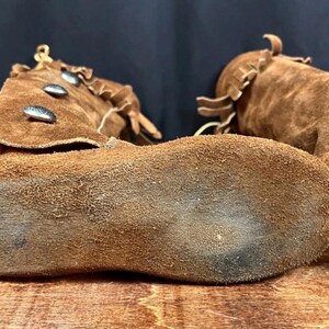 May include: Close-up of two brown suede moccasin boots. The boots have a soft, textured appearance with visible stitching and decorative elements. The soles are worn, showing signs of use. The boots are likely handmade.