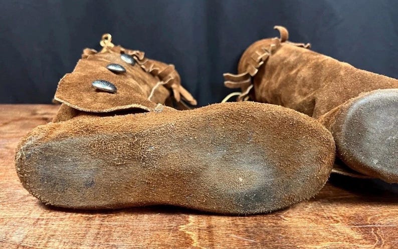 May include: Close-up of two brown suede moccasin boots. The boots have a soft, textured appearance with visible stitching and decorative elements. The soles are worn, showing signs of use. The boots are likely handmade.