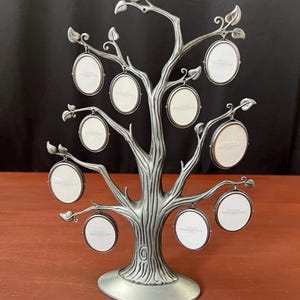 May include: A silver-toned metal tree-shaped photo display stand featuring a textured trunk and branches. The stand includes nine hanging oval-shaped picture frames, each containing a white insert with the text Things Remembered printed on it.