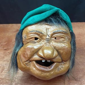 May include: A wooden mask carved to resemble an elderly person with a smiling expression. The mask features a green fabric cap, grey hair, and a detailed, aged face with wrinkles. The mouth is open, showing teeth.