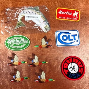May include: A selection of embroidered patches with outdoor and hunting themes. Includes a trout, flying ducks, and logos for Remington, Marlin, Colt, and Taurus. The patches are in various shapes and colours, suitable for jackets or bags.