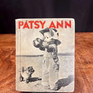 Puede incluir: Libro vintage titulado "PATSY ANN" en letras rojas. La portada presenta una imagen en blanco y negro de un niño con peto sosteniendo una pelota, con un perro pequeño cerca. El libro tiene un aspecto desgastado y envejecido.